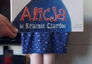 Praca Amelii Antoszewskiej - Zdjęcie przedstawia dziewczynkę ubraną w niebieską sukienkę. Do zdjęcia posłużyła książka „Alicja w krainie czarów” Lewis Carroll.